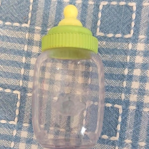 May include: A clear baby bottle with a yellow nipple and a green screw-on cap. The bottle has a flower-shaped design on the front. The bottle is empty and sits on a blue and white checkered surface.