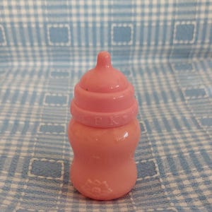 May include: A small, pink baby bottle with a nipple and a screw-on cap. The cap has the letters "CPK" printed on it. The bottle has a rounded shape and a floral design at the base. The bottle is sitting on a blue and white checkered surface.