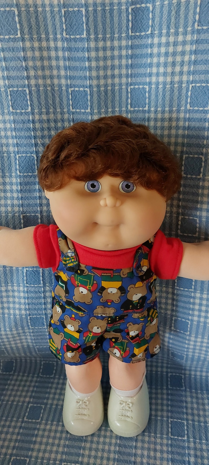 K&ouml;nnte beinhalten: Eine Cabbage Patch Kid Puppe mit braunen Haaren, blauen Augen und einem roten Shirt. Die Puppe tr&auml;gt einen blauen Overall mit einem Muster aus Teddyb&auml;ren. Die Puppe tr&auml;gt wei&szlig;e Schuhe.