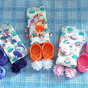Peut inclure: Trois ensembles d'accessoires pour poupée, chacun comprenant une couche à motifs, une tétine sur une chaîne de perles colorées, une paire de chaussures unies et deux pinces à cheveux en tissu en forme de fleur, déclinés en violet, orange et rose.