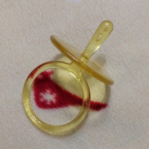 Peut inclure: Une sucette jaune transparente avec une poignée ronde et un bouclier plat et circulaire. La sucette est faite d'un matériau translucide. Le fond est une surface blanche texturée avec un motif rouge et blanc.