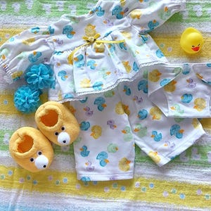 Peut inclure: Un ensemble haut à manches longues et pantalon blanc avec un motif répétitif de canards jaunes, bleus et violets, orné d'un ruban jaune et d'une petite fleur, accompagné de chaussons jaunes en forme d'ours, d'une culotte et d'un canard en caoutchouc.