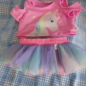 Könnte beinhalten: Rosa-violettes Einhorn-Outfit für ein Stofftier. Das Outfit besteht aus einem rosa T-Shirt mit kurzen Ärmeln und einem Einhorn-Motiv sowie einem regenbogenfarbenen Tüllrock.