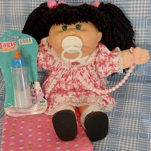 Könnte beinhalten: Eine Puppe mit schwarzem Wollhaar in Zöpfen, grünen Augen und weißem Schnuller, bekleidet mit einem rosa-weißen Blumenkleid mit Spitzenbesatz, weißen Socken und schwarzen Schuhen. Eine Magic Milk Flasche in Verpackung und ein rosa Stoffquadrat.