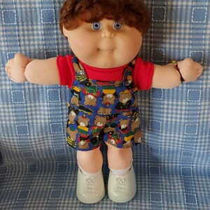 K&ouml;nnte beinhalten: Eine Cabbage Patch Kid Puppe mit braunen Haaren, blauen Augen und einem roten Shirt. Die Puppe tr&auml;gt einen blauen Overall mit einem B&auml;renmuster und tr&auml;gt ein Armband am Handgelenk. Die Puppe sitzt auf einer blau-wei&szlig; karierten Decke.