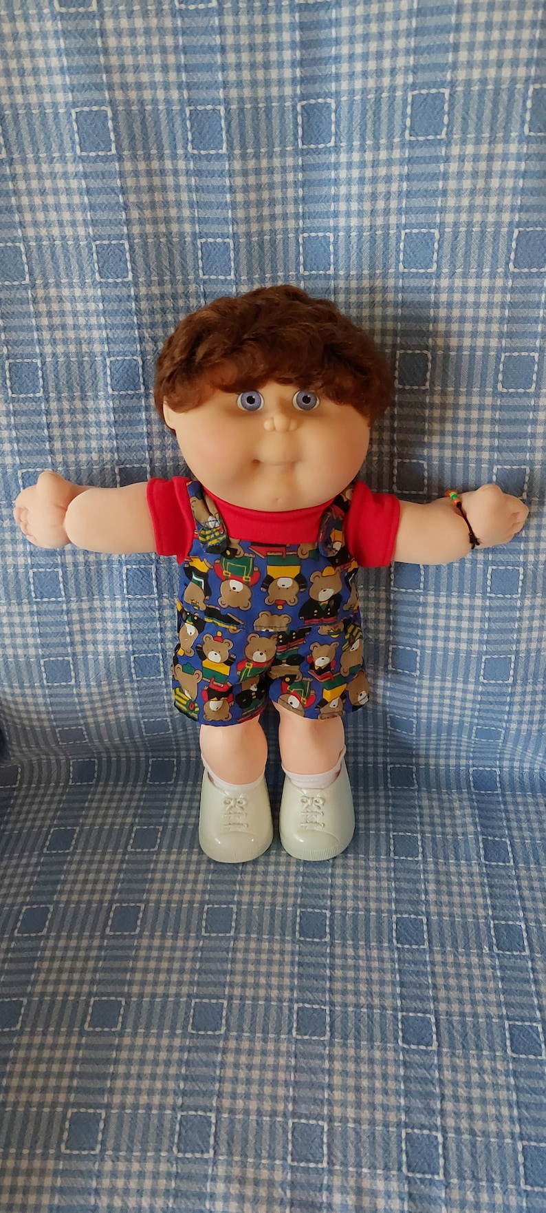 K&ouml;nnte beinhalten: Eine Cabbage Patch Kid Puppe mit braunen Haaren, blauen Augen und einem roten Shirt. Die Puppe tr&auml;gt einen blauen Overall mit einem B&auml;renmuster und tr&auml;gt ein Armband am Handgelenk. Die Puppe sitzt auf einer blau-wei&szlig; karierten Decke.