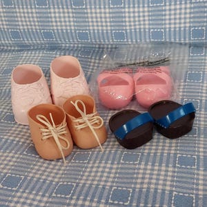 Può includere: Una collezione di scarpe da bambola in miniatura in vari stili e colori. Include due paia di scarpe da ginnastica rosa, due paia di scarpe stringate color cuoio, due paia di scarpe rosa senza lacci e due paia di sandali marroni e blu. Disposte su una superficie a scacchi blu e bianca.