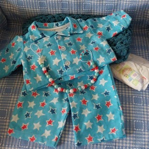 Puede incluir: Un conjunto de pijama para muñeca bebé en azul claro con un estampado de estrellas en rojo, blanco y azul marino. El conjunto incluye una camisa y un pantalón. También se incluye un clip para chupete con cuentas y un pañal.