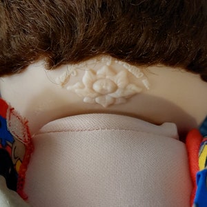 K&ouml;nnte beinhalten: Eine Cabbage Patch Kid Puppe mit braunen Haaren, die ein blaues und rotes Shirt mit einem B&auml;renmuster tr&auml;gt. Die Puppe hat eine wei&szlig;e Blume auf der R&uuml;ckseite des Nackens.