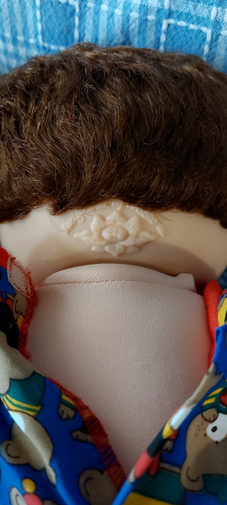 K&ouml;nnte beinhalten: Eine Cabbage Patch Kid Puppe mit braunen Haaren, die ein blaues und rotes Shirt mit einem B&auml;renmuster tr&auml;gt. Die Puppe hat eine wei&szlig;e Blume auf der R&uuml;ckseite des Nackens.