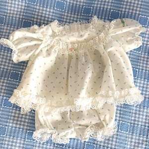 Puede incluir: Un conjunto blanco de dos piezas que incluye una blusa de manga corta y pololos a juego con un estampado de pequeñas hojas azules. La blusa tiene un cuello y dobladillo de encaje con volantes, y un pequeño detalle de rosa en el escote.