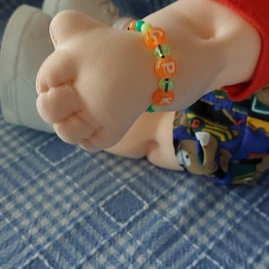 K&ouml;nnte beinhalten: Nahaufnahme eines Puppenarms, der ein buntes Perlenarmband mit den Buchstaben "CPK" tr&auml;gt.