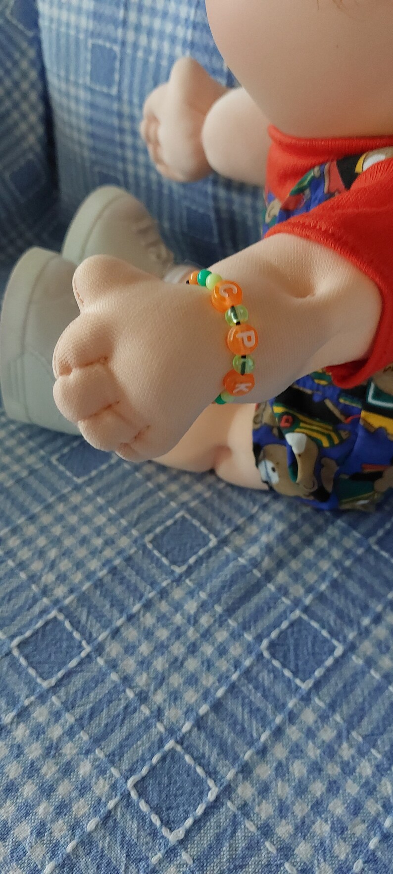 K&ouml;nnte beinhalten: Nahaufnahme eines Puppenarms, der ein buntes Perlenarmband mit den Buchstaben "CPK" tr&auml;gt.