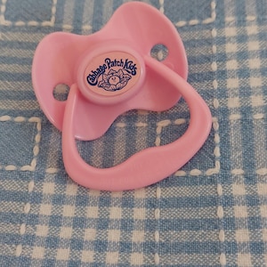May include: A pink Cabbage Patch Kids pacifier with a rounded shield and a curved handle. The pacifier has the Cabbage Patch Kids logo in a blue and white circle. The background is a blue and white checkered pattern.