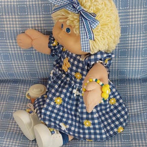 May include: A vintage doll with blonde yarn hair and blue eyes, dressed in a blue and white gingham dress with yellow flower details. The doll has a matching bow and a yellow flower bracelet. It is sitting on a blue gingham patterned surface.