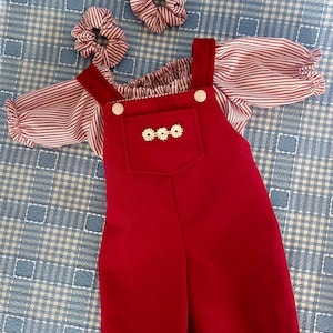 May include: A red overall set featuring a chest pocket with three small white flower embellishments and two white buttons on the straps. The set includes a red and white striped blouse with puffed sleeves and two matching red and white striped scrunchies.
