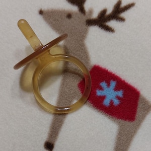 Puede incluir: Un chupete transparente de color ámbar con un anillo redondo y una tetina. El chupete está sobre una manta blanca con un diseño de reno. El reno tiene cuernos marrones, una manta roja con un copo de nieve azul.