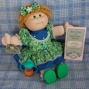 May include: A Cabbage Patch Kids doll with green eyes, wearing a green and blue patterned dress, a matching bow, and green shoes. The doll holds a bottle and a birth certificate that reads "JANE EILEEN".