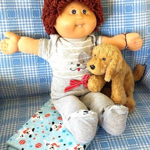 May include: A doll with brown yarn hair and a single tooth, wearing a gray striped onesie with a dog graphic and a red ribbon, holding a tan plush dog toy. The doll sits on a light blue blanket featuring a repeating pattern of white dogs and red balls.