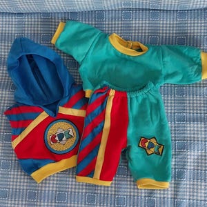 Könnte beinhalten: Ein Puppenkleidungsset bestehend aus einem blauen Kapuzenpullover mit roten und gelben Akzenten und einem runden CPK-Logo, kombiniert mit einem türkisfarbenen Langarmshirt mit gelbem Saum sowie einer türkisfarbenen Hose mit roten und blauen Streifen.