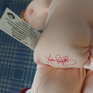 K&ouml;nnte beinhalten: Eine rosafarbene Cabbage Patch Kids Puppe mit braunen Haaren. Die Puppe tr&auml;gt einen wei&szlig;en Strampler mit einer roten, blauen und gelben gemusterten Windel. Das Etikett der Puppe lautet "31910/31911 KIDS&trade; UNDER LICENSE FROM ORIGINAL APPALACHIAN ARTWORKS INC &copy;1992, 1993 ORIGINAL APPALACHIAN ARTWORKS, INC ALL RIGHTS RESERVED MADE FOR Hasbro, Inc., Pawtucket, RI 02862 USA SURFACE WASHABLE ONLY. FILLING: ALL NEW MATERIALS: 100% POLYESTER FIBER MADE IN CHINA. C-060A"