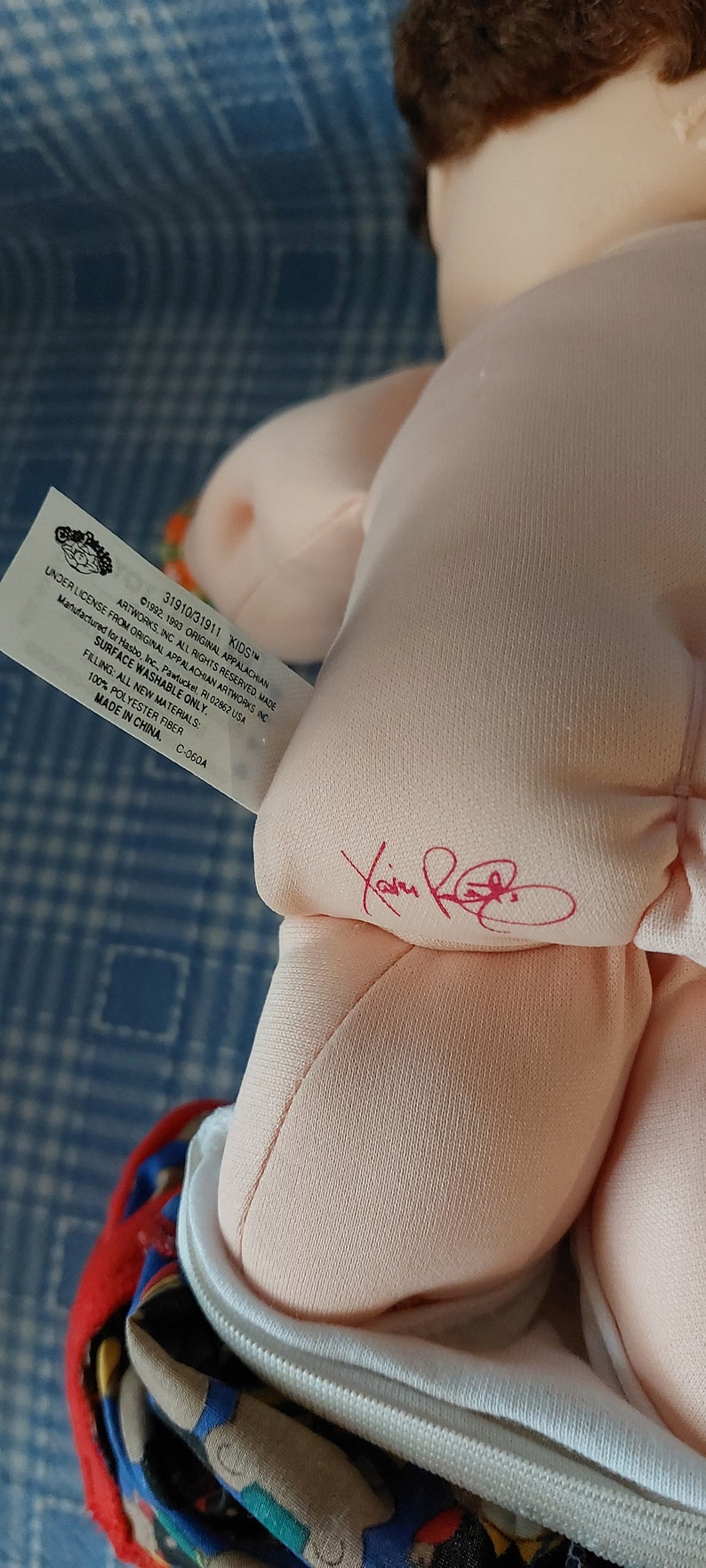 K&ouml;nnte beinhalten: Eine rosafarbene Cabbage Patch Kids Puppe mit braunen Haaren. Die Puppe tr&auml;gt einen wei&szlig;en Strampler mit einer roten, blauen und gelben gemusterten Windel. Das Etikett der Puppe lautet "31910/31911 KIDS&trade; UNDER LICENSE FROM ORIGINAL APPALACHIAN ARTWORKS INC &copy;1992, 1993 ORIGINAL APPALACHIAN ARTWORKS, INC ALL RIGHTS RESERVED MADE FOR Hasbro, Inc., Pawtucket, RI 02862 USA SURFACE WASHABLE ONLY. FILLING: ALL NEW MATERIALS: 100% POLYESTER FIBER MADE IN CHINA. C-060A"