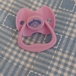 May include: A light pink Cabbage Patch Kids pacifier. The pacifier has a rounded shield with the Cabbage Patch Kids logo in a blue circle. The handle and nipple are also light pink. The pacifier is on a blue and white checkered surface.
