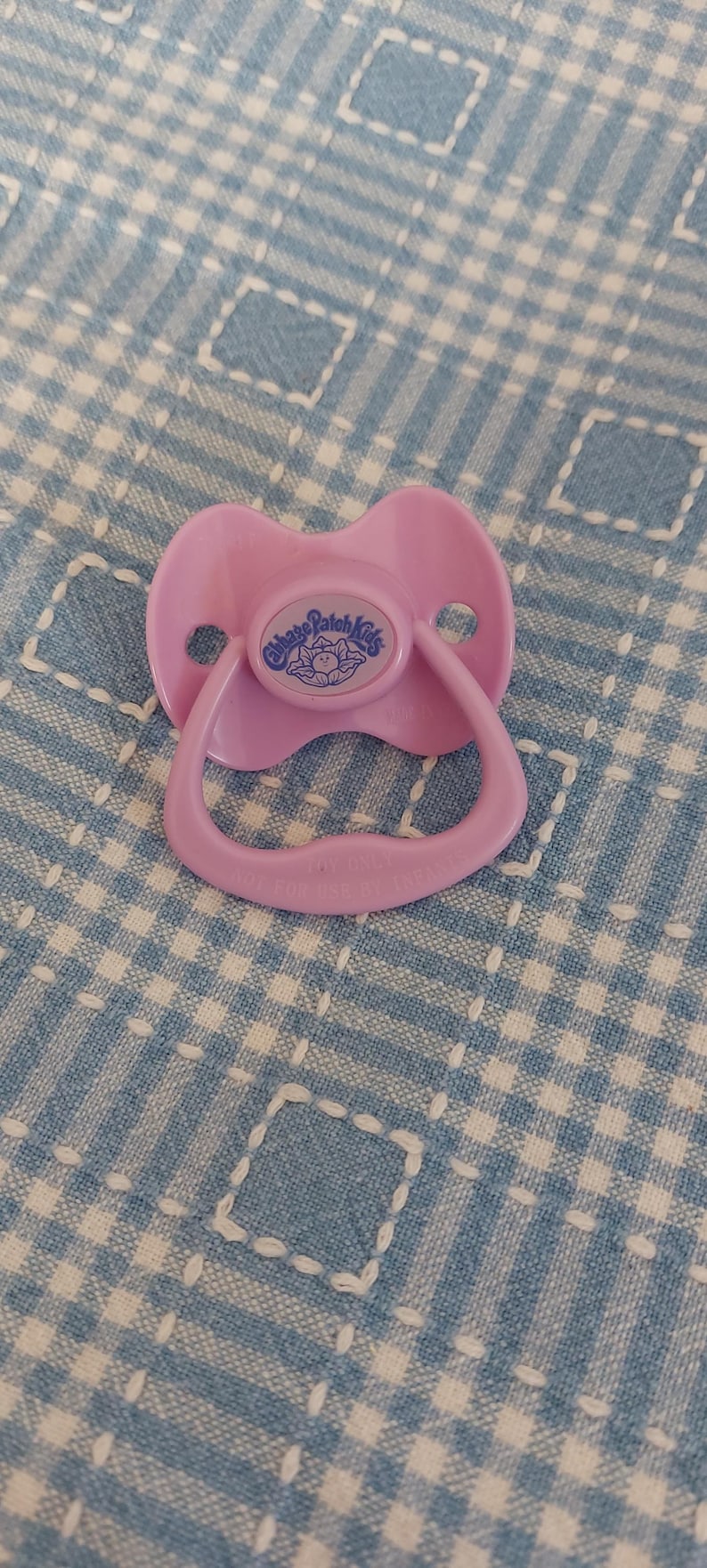K&ouml;nnte beinhalten: Ein hellrosa Cabbage Patch Kids Schnuller. Der Schnuller hat einen abgerundeten Schild mit dem Cabbage Patch Kids-Logo in einem blauen Kreis. Der Griff und der Sauger sind ebenfalls hellrosa. Der Schnuller liegt auf einer blau-wei&szlig; karierten Oberfl&auml;che.