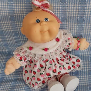 May include: A vintage doll with a bald head, blue eyes, and a smiling expression. The doll wears a white dress with a strawberry print, lace trim, and a matching bow. A bracelet with pink and green beads adorns the doll's wrist.