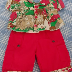May include: A baby's two-piece outfit with a patterned top and red shorts. The top has a colourful print with green, red, and brown accents. The shorts have two pockets and a decorative trim at the hem. The outfit is designed for a baby.