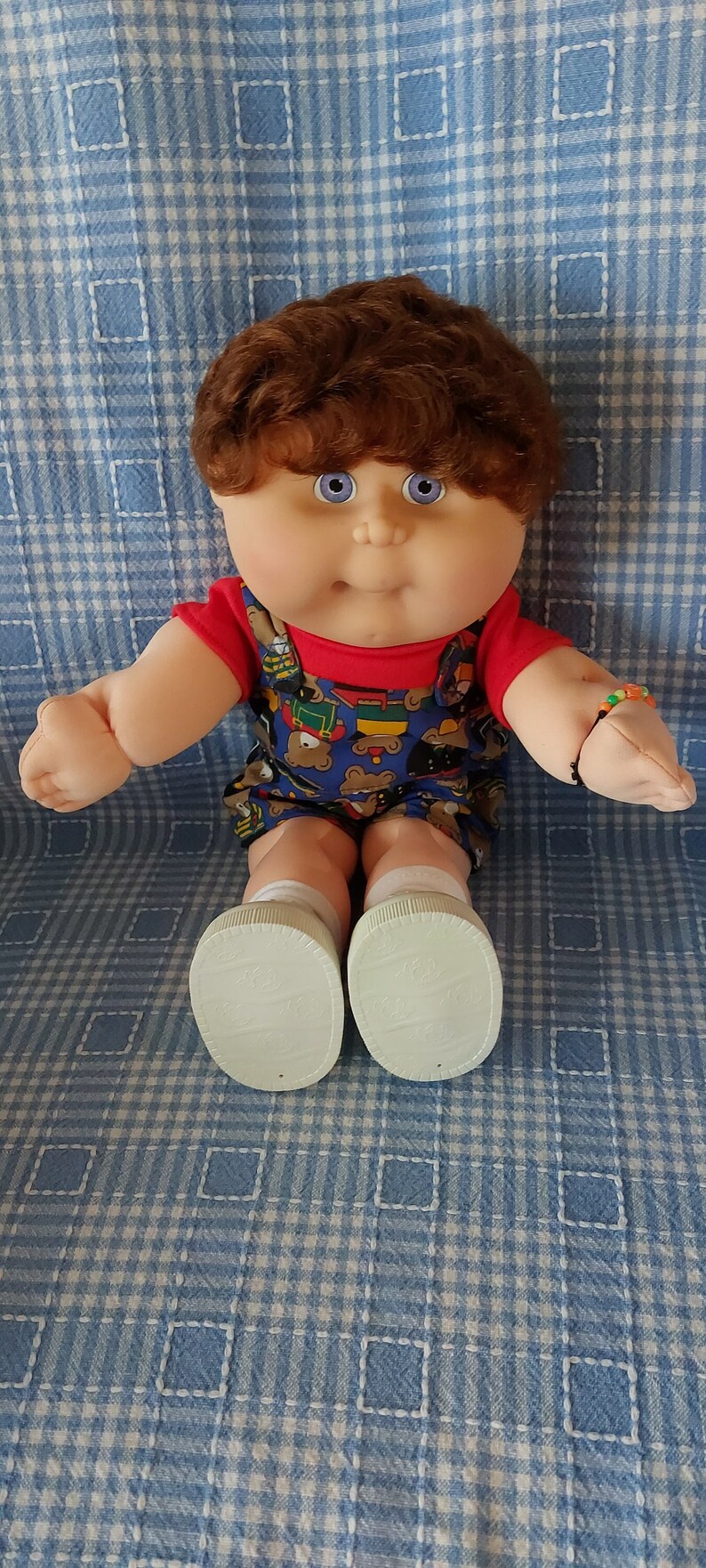 K&ouml;nnte beinhalten: Eine Cabbage Patch Kid Puppe mit braunen Haaren, blauen Augen, einem roten Shirt und einer blauen und gelben gemusterten Latzhose. Die Puppe tr&auml;gt wei&szlig;e Schuhe und ein Armband am Handgelenk.