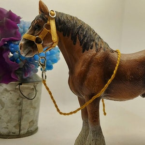 May include: A brown and white horse figurine with a yellow halter and lead rope. The horse stands next to a galvanized metal bucket filled with purple and blue flowers. The background is white.