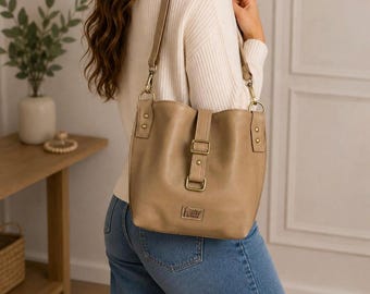 May include: A tan bucket-style shoulder bag featuring a vertical center strap with a rectangular metal buckle, gold-toned hardware, and a small rectangular brand label at the bottom. The bag has an adjustable shoulder strap attached with metal clips.