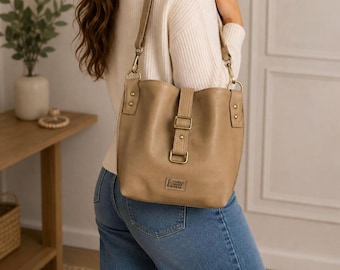 May include: A tan bucket-style shoulder bag featuring a vertical strap with a rectangular buckle closure, antique brass-toned metal hardware, and a small rectangular brand label at the bottom center. The bag has a smooth finish and a long shoulder strap.