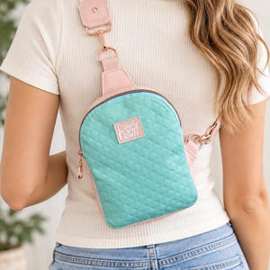 May include: A quilted teal crossbody sling bag with light pink trim and rose gold hardware. The bag features a rectangular patch with the text "calm your knits" and a wide, adjustable strap with a rose gold clip attachment and pink accents.