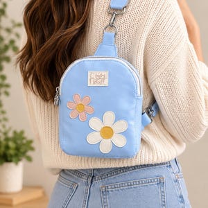 May include: A light blue crossbody sling bag featuring a rectangular shape with rounded top corners, a silver zipper, and two flower appliqués in pink and white with yellow centers. A small rectangular patch displays the text calm your tits.