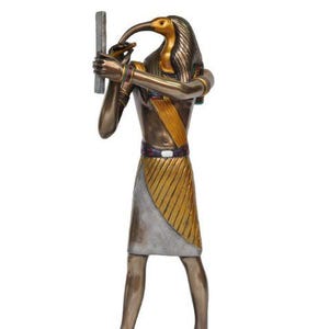 Puede incluir: Estatua de bronce del dios egipcio Thot, representado con cabeza de ibis. La figura sostiene un báculo y viste una prenda dorada y beige. La estatua se alza sobre una base rectangular.