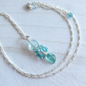 May include: A silver necklace featuring a pendant with light blue and turquoise gemstones. The pendant has a cluster of small round stones and two larger, teardrop-shaped stones. The necklace chain is a delicate, double strand design.