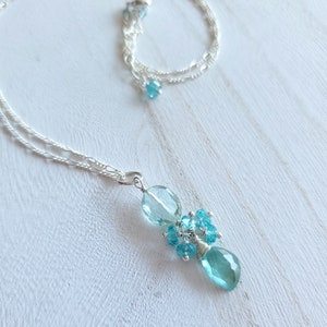 May include: A delicate silver necklace with a cluster of light blue gemstones. The pendant features a large teardrop-shaped stone, surrounded by smaller, round blue gems. The necklace chain is a fine, twisted design.
