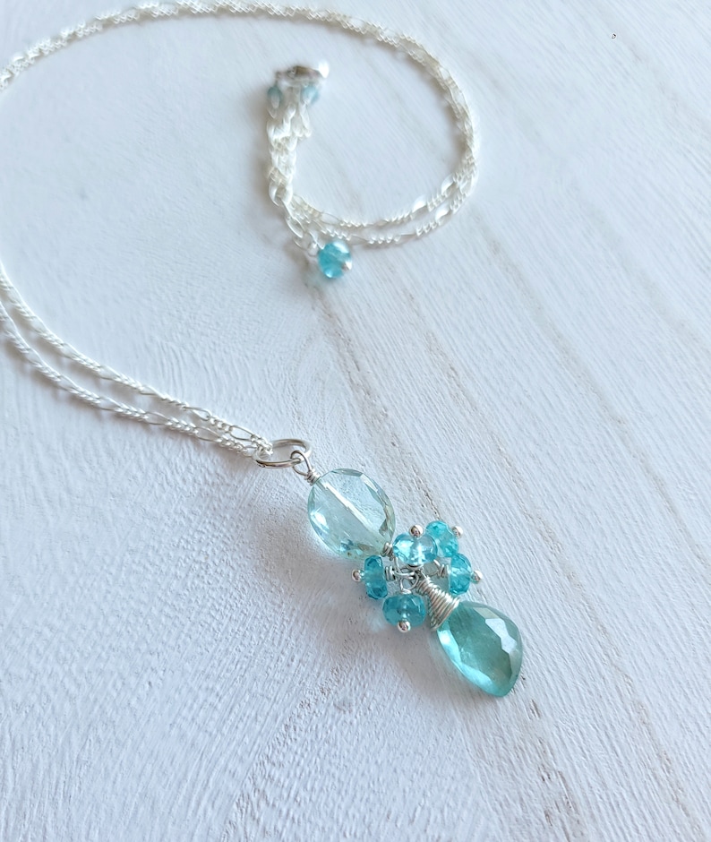 May include: A delicate silver necklace with a cluster of light blue gemstones. The pendant features a large teardrop-shaped stone, surrounded by smaller, round blue gems. The necklace chain is a fine, twisted design.