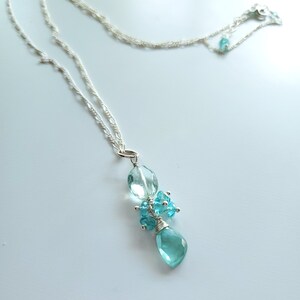May include: A delicate silver necklace featuring a cluster of light blue gemstones. The pendant includes an oval-shaped gem, a teardrop-shaped gem, and a cluster of smaller gems, all wire-wrapped. The necklace has a delicate chain.
