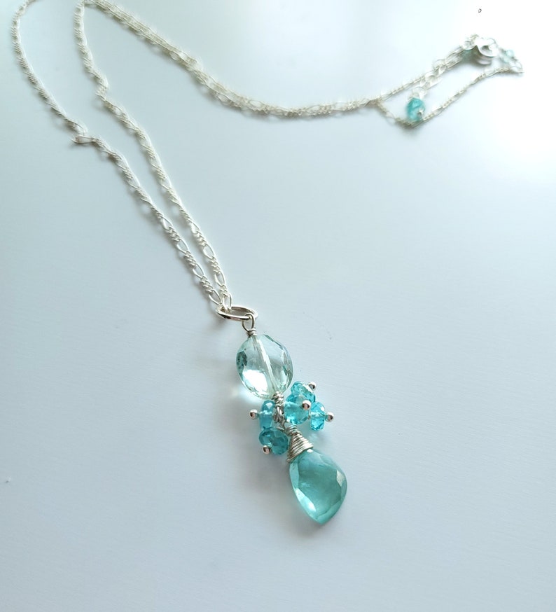 May include: A delicate silver necklace featuring a cluster of light blue gemstones. The pendant includes an oval-shaped gem, a teardrop-shaped gem, and a cluster of smaller gems, all wire-wrapped. The necklace has a delicate chain.