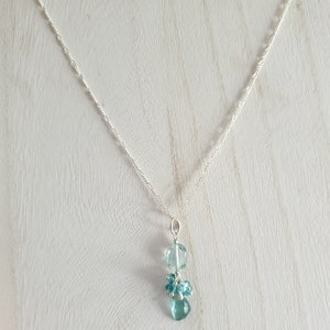 May include: A delicate silver necklace featuring a pendant with light blue, faceted beads. The pendant has a cluster of small beads and two larger, round beads. The necklace chain is thin and has a subtle, textured design.