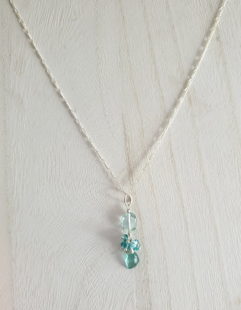 May include: A delicate silver necklace featuring a pendant with light blue, faceted beads. The pendant has a cluster of small beads and two larger, round beads. The necklace chain is thin and has a subtle, textured design.