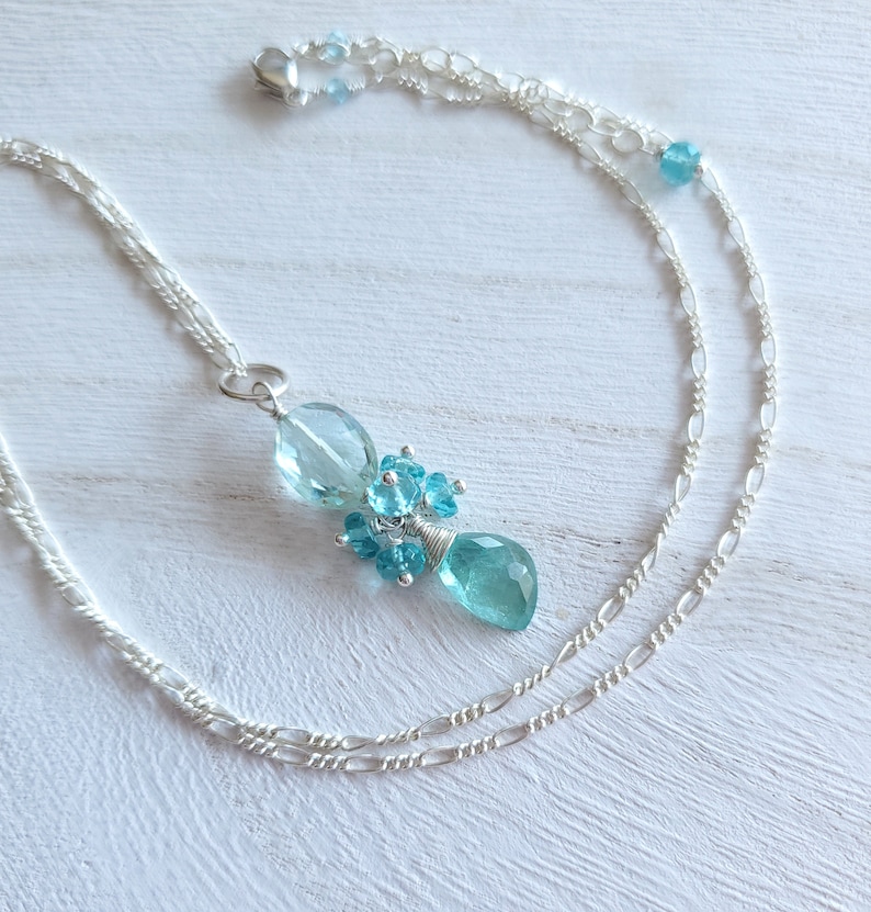 May include: A silver necklace featuring a pendant with light blue and turquoise gemstones. The pendant has a cluster of small round stones and two larger, teardrop-shaped stones. The necklace chain is a delicate, double strand design.