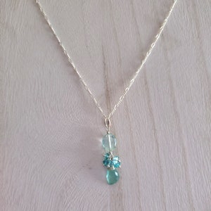 May include: A delicate silver necklace featuring a pendant with three light blue gemstone clusters. The top stone is a large, clear, rounded shape, followed by a cluster of smaller, round stones, and a teardrop-shaped stone at the bottom.