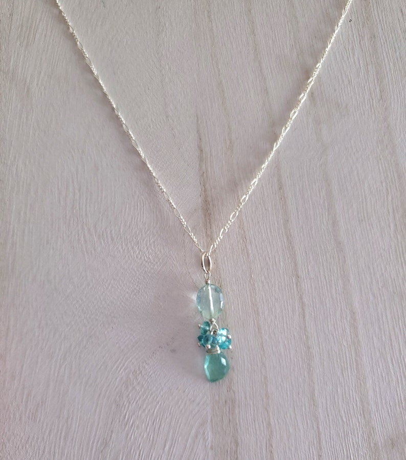 May include: A delicate silver necklace featuring a pendant with three light blue gemstone clusters. The top stone is a large, clear, rounded shape, followed by a cluster of smaller, round stones, and a teardrop-shaped stone at the bottom.