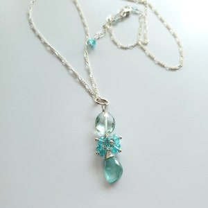 May include: A delicate silver necklace featuring a pendant with light blue gemstones. The pendant has a large, faceted teardrop-shaped stone at the bottom, with smaller blue stones clustered above. The necklace chain is a fine, linked design.