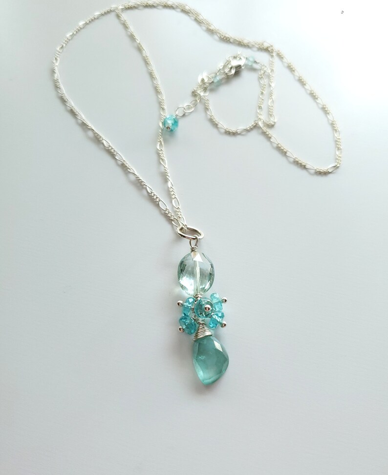 May include: A delicate silver necklace featuring a pendant with light blue gemstones. The pendant has a large, faceted teardrop-shaped stone at the bottom, with smaller blue stones clustered above. The necklace chain is a fine, linked design.