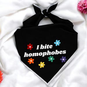 May include: Black dog bandana with white trim and the text "I bite homophobes" in white, surrounded by colorful flower graphics. The bandana is knotted at the top and rests on a white surface with colorful flowers.